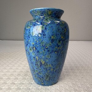 May include: A ceramic vase with a glossy finish, featuring a vibrant blue base color speckled with dark blue and yellow crystalline patterns. The vase has a rounded body, a narrow neck, and a flared rim, showcasing a smooth, reflective surface.