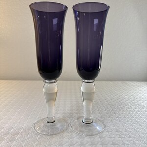 May include: Two tall champagne flutes featuring deep purple glass bowls and clear glass stems with circular bases. The flutes have a smooth, elongated silhouette with a flared rim, showcasing a distinct contrast between the dark purple upper section and the transparent lower stem and base.