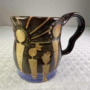 May include: A ceramic mug with a dark brown glaze featuring a light tan abstract design of four figures standing under a sunburst pattern. The mug has a curved handle and a distinct blue band around the base, showcasing a hand-crafted aesthetic.