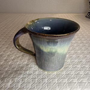 May include: A ceramic mug featuring a flared rim and a curved handle. The exterior and interior display a blended glaze of deep blue, slate gray, and pale yellow, with a dark brown rim and handle edge, creating a smooth, glossy, multi-toned finish.