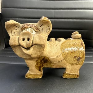 May include: A tan ceramic piggy bank with a textured, matte finish. The pig features carved details on the ears and face, a prominent snout, and a small curled tail. Darker brown glazed patches accent the legs and rear, with a slot on the back.