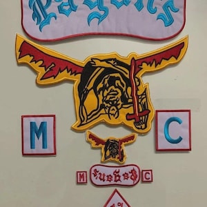 May include: A collection of embroidered patches including a large white rocker patch with the blue text Pagan's, a large yellow and red winged figure patch, two square patches with blue letters M and C, a small winged figure patch, a small Pagan's patch, and a diamond patch with 1% text.