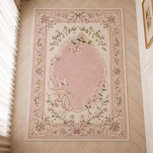 Può includere: Un tappeto rettangolare caratterizzato da un medaglione ovale centrale rosa tenue circondato da intricati ghirlande floreali, nastri e motivi botanici nei toni del verde, crema e rosa antico, incorniciato da un bordo decorativo con accenti floreali.