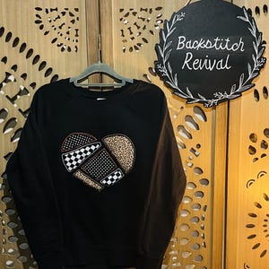Puede incluir: Sudadera negra con un diseño de corazón de patchwork. El corazón presenta una mezcla de patrones, incluyendo cuadros, lunares y estampado de leopardo. Un letrero redondo negro con el texto "Backstitch Revival" cuelga cerca.