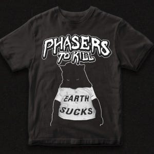 May include: Black t-shirt with the text "PHASERS TO KILL" in white, above a cartoon figure wearing a shirt that says "EARTH SUCKS". The graphic tee has a crew neck and short sleeves.