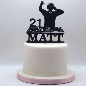 May include: A black cake topper featuring a silhouette of a DJ with headphones at a mixing console, the number 21, and the name MATT in capital letters, positioned on top of a smooth white cake with a light pink base trim.