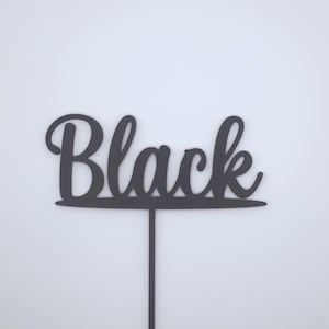 May include: A black cake topper featuring the word Black written in a cursive script font. The lettering is connected to a horizontal line that sits atop a single vertical stake, designed for insertion into a cake or other food item.