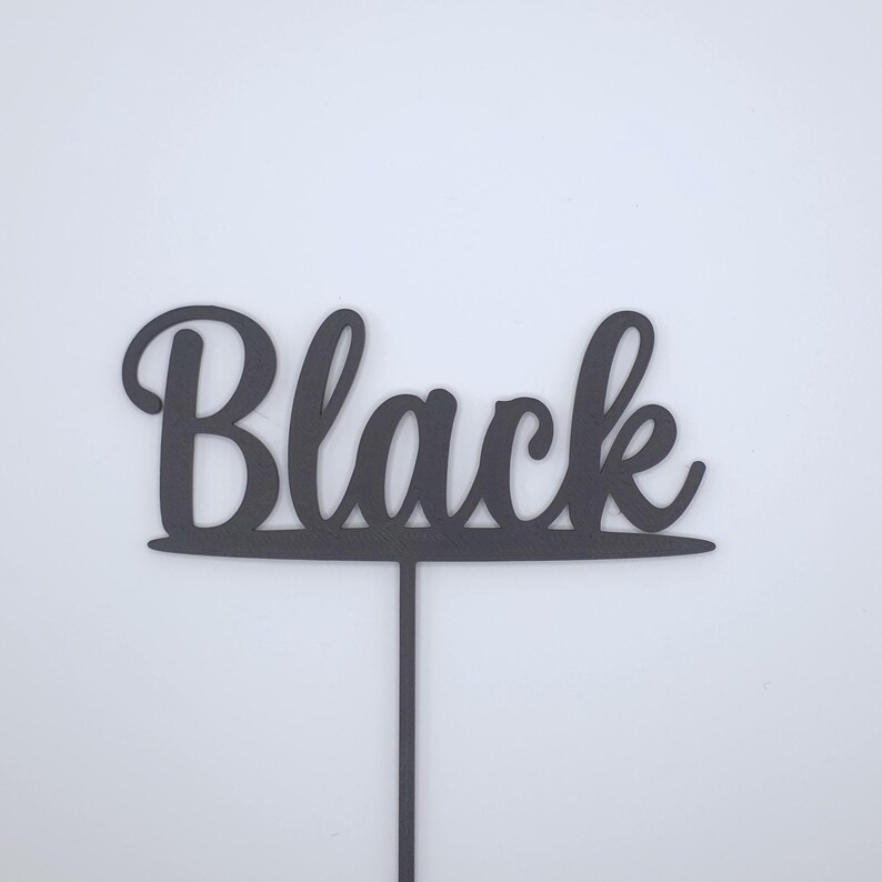 May include: A black cake topper featuring the word Black written in a cursive script font. The lettering is connected to a horizontal line that sits atop a single vertical stake, designed for insertion into a cake or other food item.