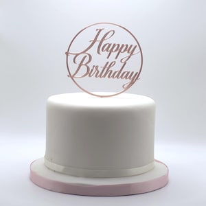 May include: A round white cake with a pink base, topped with a rose gold "Happy Birthday" cake topper. The topper features cursive lettering within a circular frame. The cake is a classic design.