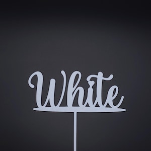 May include: A white cake topper featuring the word White in a cursive script font. The lettering is connected to a horizontal base line with a single vertical stake extending downward from the centre, designed for insertion into a cake or food item.