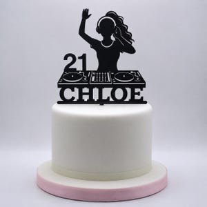 May include: A black cake topper featuring a silhouette of a DJ wearing headphones with one hand raised. The design includes the number 21, a mixing console with two turntables, and the name CHLOE in bold capital letters, all atop a white cake.