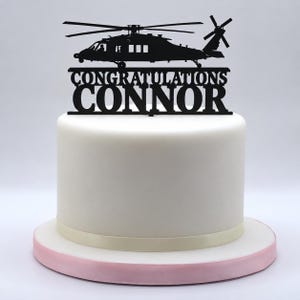 May include: A black cake topper featuring a silhouette of a military helicopter above the text CONGRATULATIONS CONNOR. The topper is inserted into a smooth white cylindrical cake with a light pink base and a thin ribbon around the bottom.