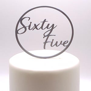 Könnte beinhalten: Ein grauer Tortenaufsatz mit den Worten "Sixty Five" in kursiver Schrift, in einem kreisförmigen Rahmen. Der Aufsatz befindet sich auf einem weiß glasierten Kuchen, ideal für einen Geburtstag.