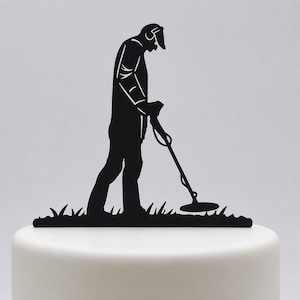 May include: A black silhouette cake topper featuring a person wearing a cap and headphones while using a metal detector. The figure is positioned over a base of stylised grass, designed to be inserted into the top of a cake for decoration.