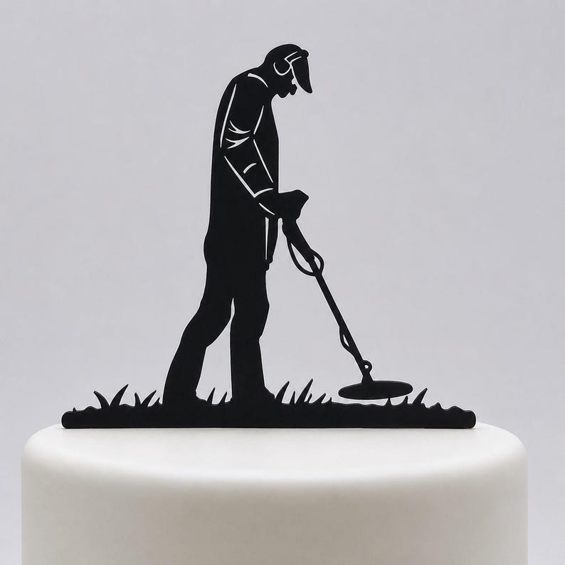 May include: A black silhouette cake topper featuring a person wearing a cap and headphones while using a metal detector. The figure is positioned over a base of stylised grass, designed to be inserted into the top of a cake for decoration.
