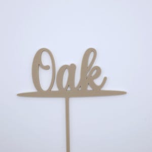 May include: A light beige cake topper featuring the word Oak in a cursive script font. The letters are connected to a horizontal line that extends across the width of the word, with a single vertical stem extending downward from the centre.