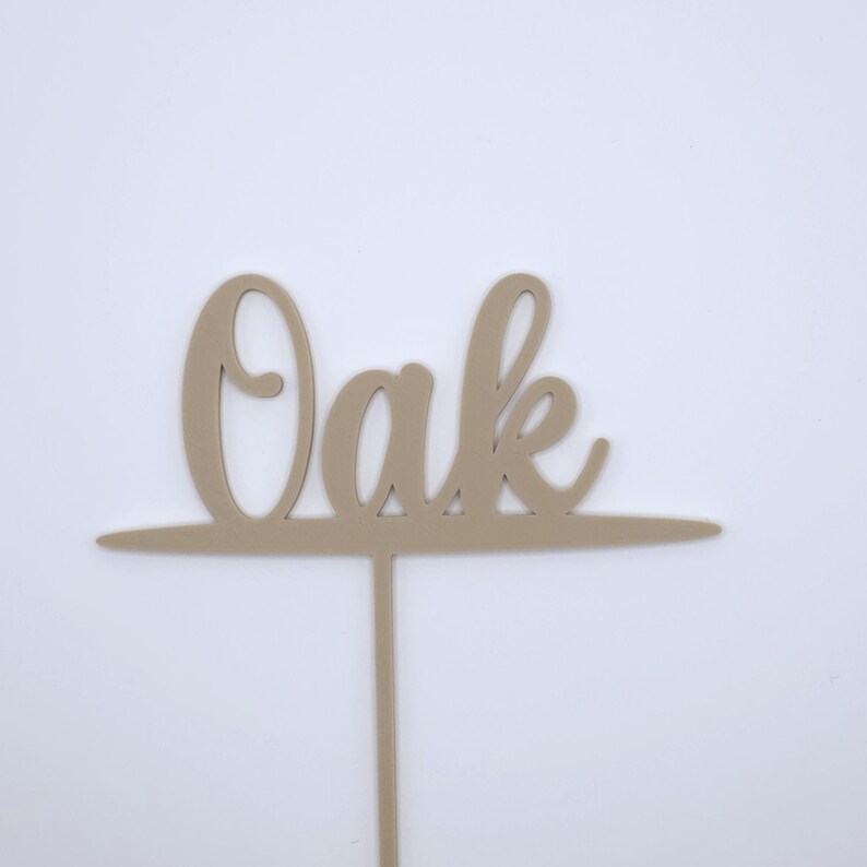 May include: A light beige cake topper featuring the word Oak in a cursive script font. The letters are connected to a horizontal line that extends across the width of the word, with a single vertical stem extending downward from the centre.