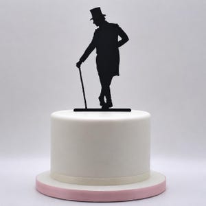 Op de afbeelding: Een zwarte silhouet taarttopper van een persoon met een hoge hoed en een lange jas, staand met gekruiste benen en leunend op een wandelstok. De topper staat bovenop een gladde, cilindervormige witte taart met een lichtroze onderkant.