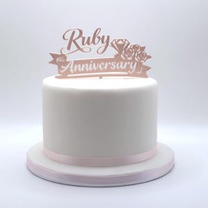 May include: A white cake with a pink ribbon around the base. A rose gold cake topper reads "Ruby Anniversary" with a diamond and floral design. The cake is on a white cake stand.