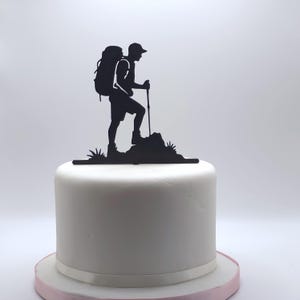 May include: A black silhouette cake topper featuring a hiker with a backpack and walking stick, standing on a rocky outcrop. The topper is placed on a white cake with a pink base, suitable for a hiking or outdoor-themed celebration.