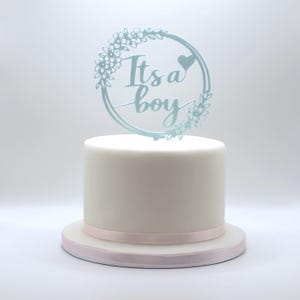Könnte beinhalten: Eine weiße Torte mit einem hellrosa Band und einem blauen Tortenaufsatz mit der Aufschrift "Its a boy". Der Tortenaufsatz hat ein florales Kranzdesign mit einem Herz.