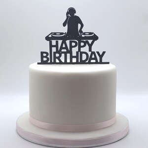 Puede incluir: Un pastel blanco liso con una cinta rosa claro alrededor de la base, coronado con un adorno negro de silueta que muestra a un DJ en una mesa de mezclas y el texto HAPPY BIRTHDAY en letras mayúsculas negritas sobre el equipo de tocadiscos.