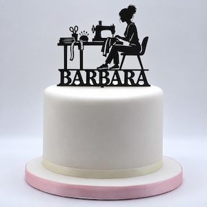 May include: A black cake topper featuring a silhouette of a person sitting at a table with a sewing machine, scissors, and a pincushion. The name BARBARA is displayed in bold capital letters at the base of the topper, which is placed on a white cake.