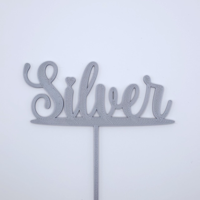 May include: A light grey cake topper featuring the word Silver in a cursive script font. The lettering is connected by a horizontal line at the base, which extends into a single vertical stake for insertion into a cake or other food item.