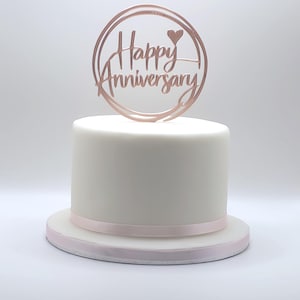 May include: A white cake with a pink ribbon around the base, topped with a rose gold circular cake topper that reads "Happy Anniversary" with a heart symbol. The cake is on a white surface.
