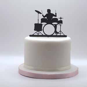 May include: A black silhouette cake topper featuring a drummer playing a drum set with a bass drum, snare, toms, and cymbals. The topper is positioned on top of a smooth, round, white frosted cake with a light pink ribbon trim around the base.
