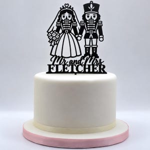 May include: A black cake topper featuring a nutcracker bride and groom silhouette, with the text Mr and Mrs FLETCHER below them, positioned on top of a smooth white cake with a light pink base and a cream-coloured ribbon trim.