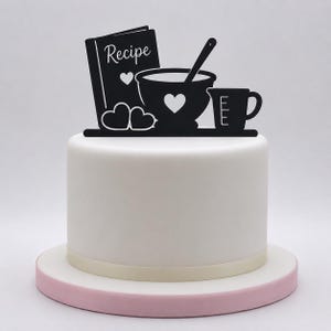 Könnte beinhalten: Ein schwarzer Tortenaufleger mit der Silhouette eines Rezeptbuchs mit dem Wort Recipe, einer Rührschüssel mit Löffel, zwei Herzen und einem Messbecher. Der Aufleger steht auf einer glatten weißen Torte mit cremefarbenem Band und hellrosa Boden.