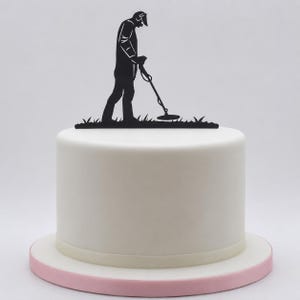 May include: A black cake topper featuring a silhouette of a person using a metal detector whilst standing on a patch of grass. The topper is placed on top of a smooth, round, white frosted cake that sits on a light pink circular cake board.