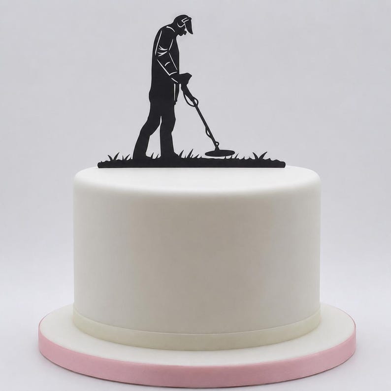 May include: A black cake topper featuring a silhouette of a person using a metal detector whilst standing on a patch of grass. The topper is placed on top of a smooth, round, white frosted cake that sits on a light pink circular cake board.
