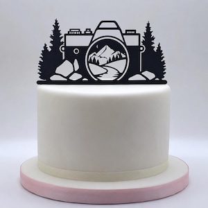 Puede incluir: Un adorno para pastel negro con la silueta de una cámara que muestra un paisaje de montaña y bosque dentro del lente. El diseño incluye pinos a ambos lados de la cámara y se coloca sobre un pastel blanco cilíndrico con una base rosa.