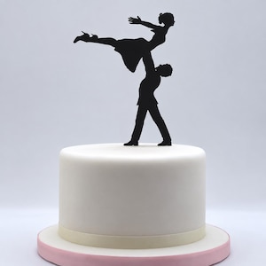 Peut inclure: Une décoration de gâteau en silhouette noire représentant deux personnages dans une pose de danse, l'un soulevant l'autre dans les airs. La décoration est placée sur un gâteau rond, lisse et glacé de blanc avec une bordure de base claire.