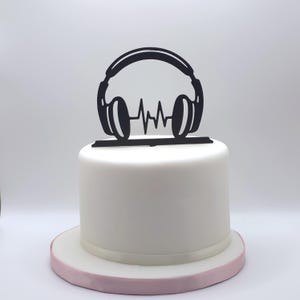Puede incluir: Un adorno para pastel negro que presenta la silueta de unos auriculares con un gráfico de onda sonora central, colocado sobre un pastel blanco cilíndrico y liso que descansa sobre una base redonda con un borde rosa claro.