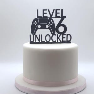 May include: A black cake topper featuring a video game controller silhouette, the number 6, and the text LEVEL UNLOCKED, positioned on top of a smooth white cylindrical cake with a light pink ribbon trim around the base of the cake.