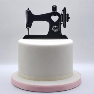 May include: A black silhouette of a sewing machine cake topper featuring a heart-shaped cutout and a circular floral emblem, positioned atop a smooth white cylindrical cake with a light pink base, creating a clean and focused visual presentation.