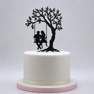 May include: A black cake topper silhouette featuring a large tree with a swing hanging from a branch. Two figures are seated together on the swing. The topper is positioned on top of a smooth, cylindrical white cake with a light pink base layer.