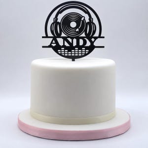 Könnte beinhalten: Ein schwarzer Tortenstecker mit einem kreisförmigen Design, das Kopfhörer, eine Schallplatte und einen Equalizer zeigt. Der Name ANDY steht in der Mitte des Steckers. Der Tortenstecker befindet sich auf einer glatten, weißen, zylindrischen Torte.