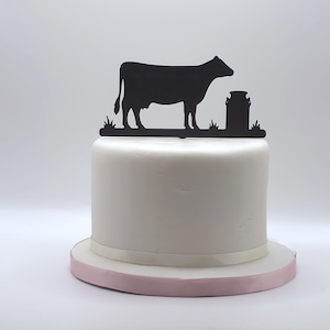 May include: A black silhouette cake topper featuring a cow standing next to a milk churn on a base of grass. The topper is positioned on top of a smooth, round, white frosted cake that sits on a circular base with a light pink ribbon trim.