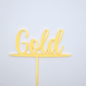 May include: A gold-coloured cake topper featuring the word Gold written in a cursive script font. The lettering is connected to a horizontal line at the base, which extends into a single vertical stake for insertion into a cake or other food item.