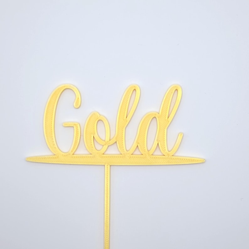 May include: A gold-coloured cake topper featuring the word Gold written in a cursive script font. The lettering is connected to a horizontal line at the base, which extends into a single vertical stake for insertion into a cake or other food item.