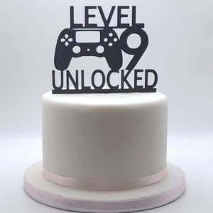 Pode incluir: Um bolo branco redondo com uma fita rosa. Um topo de bolo preto com um design de controlador e as palavras "LEVEL UNLOCKED 9". Um bolo comemorativo, perfeito para um aniversário ou evento de jogos.