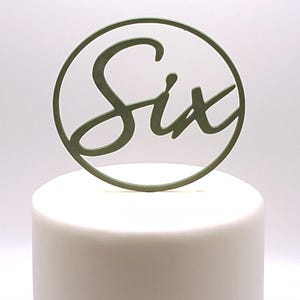 Könnte beinhalten: Ein salbeigrüner Tortenaufsatz mit dem Wort "Six" in einer Schreibschrift, umgeben von einem passenden grünen Ring. Der Aufsatz befindet sich auf einem weiß glasierten Kuchen und erzeugt ein schlichtes, aber elegantes Design.
