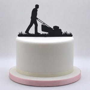 Op de afbeelding: Een witte taart met een zwarte silhouet taarttopper van een persoon die een grasmaaier over een stuk gras duwt. De taart staat op een ronde ondergrond met een lichtroze rand en een gladde witte afwerking aan de zijkanten en bovenkant.