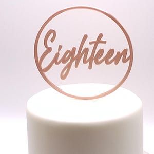 Może przedstawiać: Różowo-złoty topper na tort z napisem "Eighteen" w ozdobnej czcionce, otoczony pasującym różowo-złotym okręgiem. Topper umieszczony na białym, lukrowanym torcie, idealny na 18. urodziny.