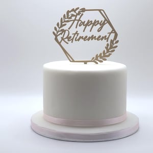 Może przedstawiać: Białe ciasto z różową wstążką u podstawy. Złoty topper na ciasto w kształcie sześciokąta z napisem "Happy Retirement" i akcentami liści umieszczony na wierzchu. Ciasto stoi na białej powierzchni.