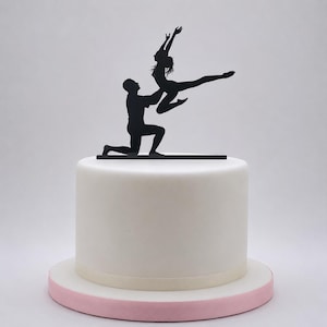 Peut inclure: Une décoration de gâteau en silhouette noire représentant un personnage agenouillé sur une base horizontale, soulevant un autre personnage dans une pose de danse dynamique. Le tout est posé sur un gâteau blanc cylindrique à bordure rose.